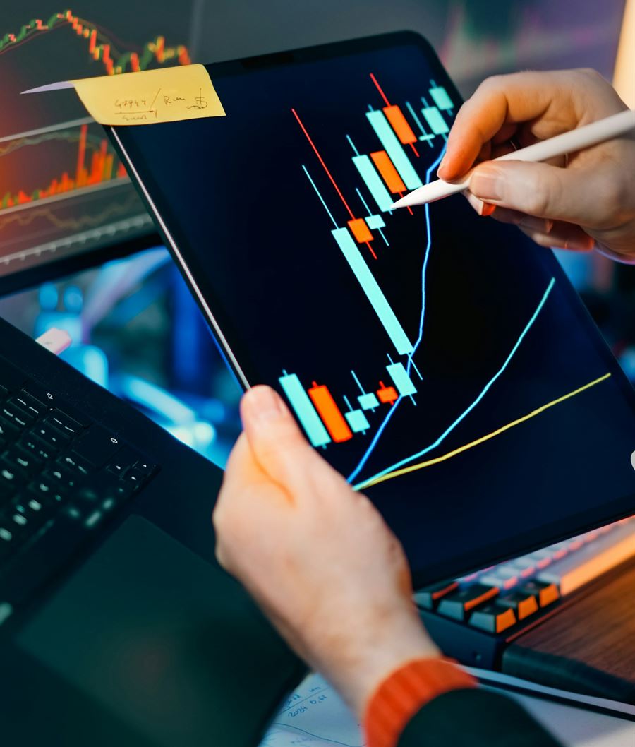 Hands marking a candlestick chart on a tablet, with more charts on the screens behind